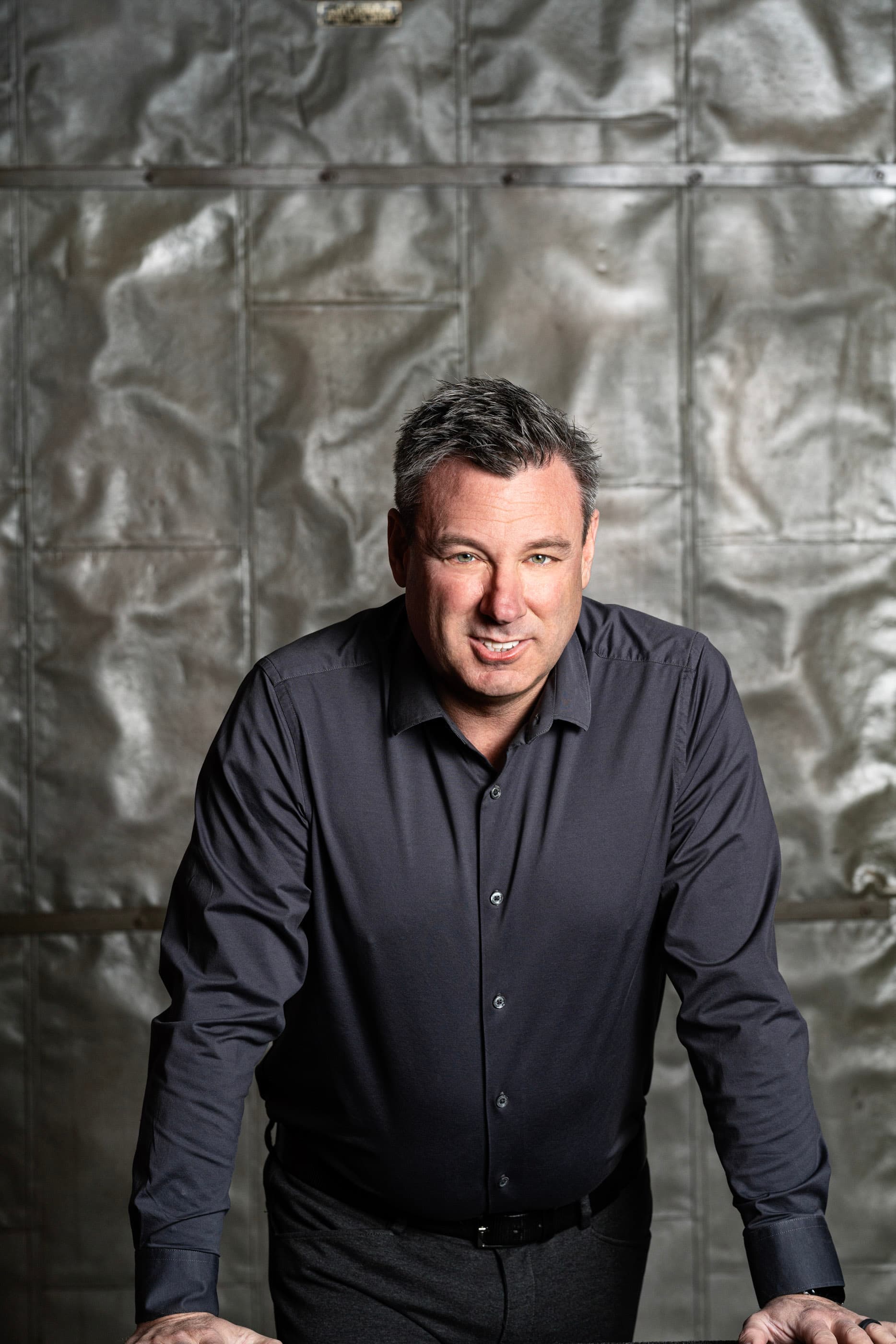 Smiling man in a dark grey shirt leaning forward against a textured metal wall.